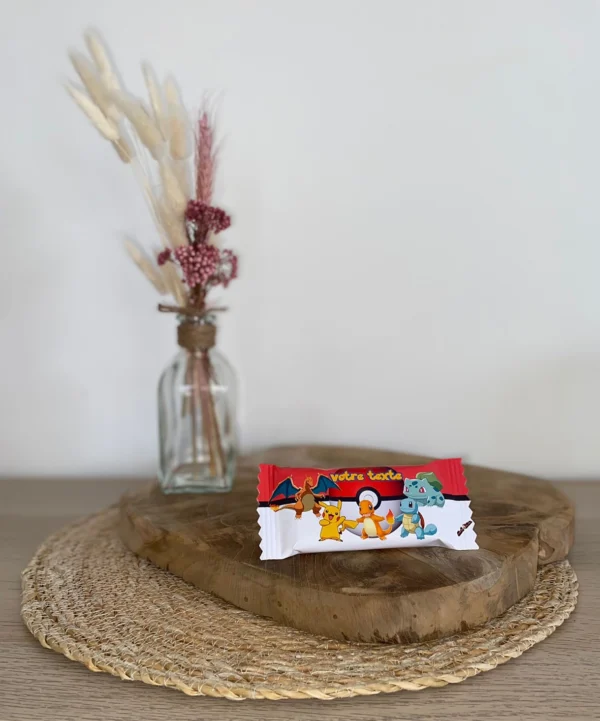 Barre Kinder Bueno personnalisée avec un emballage Pokémon, posée sur un support en bois avec une décoration florale en arrière-plan.